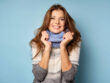 A beautiful brunette in a sweater and scarf stands on a blue background, smiling broadly at the frame. Horizontal photo